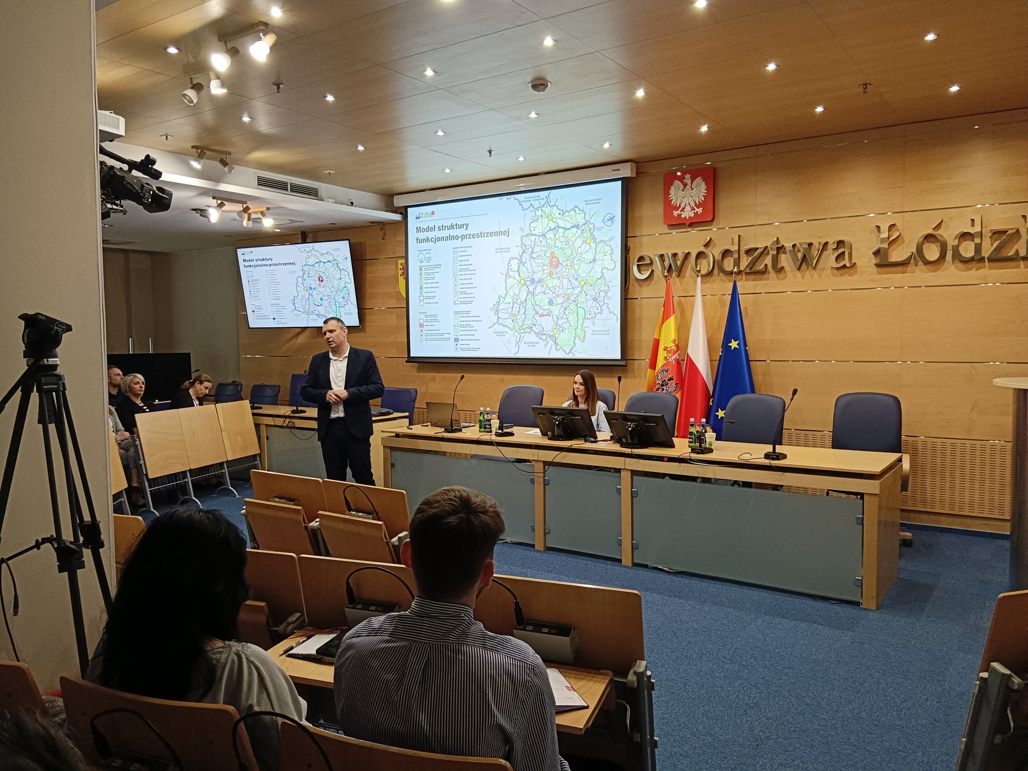 Zdjęcie z konsultacji społecznych, które odbyły się w Łodzi w sali Urzędu Marszałkowskiego, dotyczącego aktualizacji Strategii Rozwoju Województwa Łódzkiego. Na pierwszym planie zdjęcia widać tyłem siedzących uczestników spotkania. Na drugim planie zdjęcia widać: godło Polski, napis Województwo Łódzkie, poniżej flagi Urzędu Marszałkowskiego, Polski oraz Unii Europejskiej; w centralnej części widać tło rzutnika wyświetlającym slajd pt.: "Model struktury funkcjonalno-przestrzennej" oraz mapę przedstawiającą województwo łódzkie. Po lewej stronie, obok prezentacji, przodem do widowni stoi Dyrektor BPRWŁ.